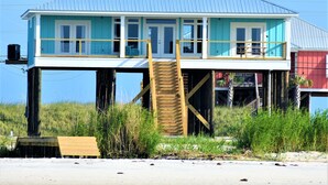 Una casa de playa con una escalera de madera que conduce a un balcón, rodeada de hierba alta y una playa arenosa.