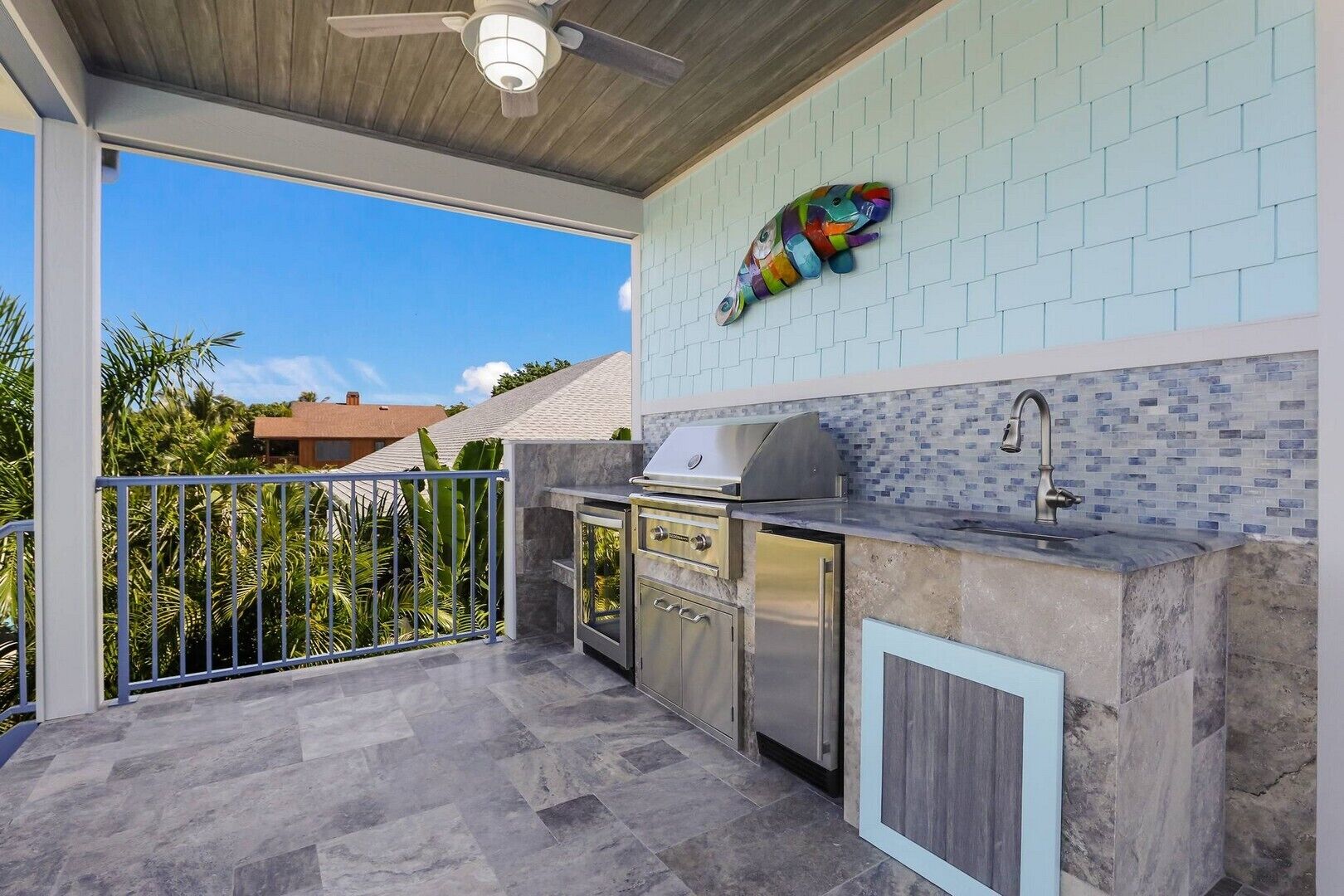 An outdoor kitchen area with a grill, sink, and storage cabinets, set against a light blue tiled wall and a metal railing.