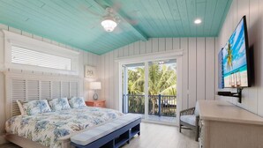A bedroom with a bed, a nightstand, a bench, a TV, and a balcony with a view of palm trees.