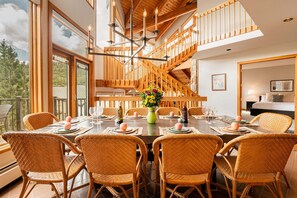 A dining area with a large wooden table set for six, surrounded by wicker chairs, a chandelier, and a staircase leading to an upper level.