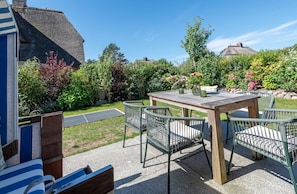 A patio with a wooden table and metal chairs, surrounded by a garden with various plants and flowers.