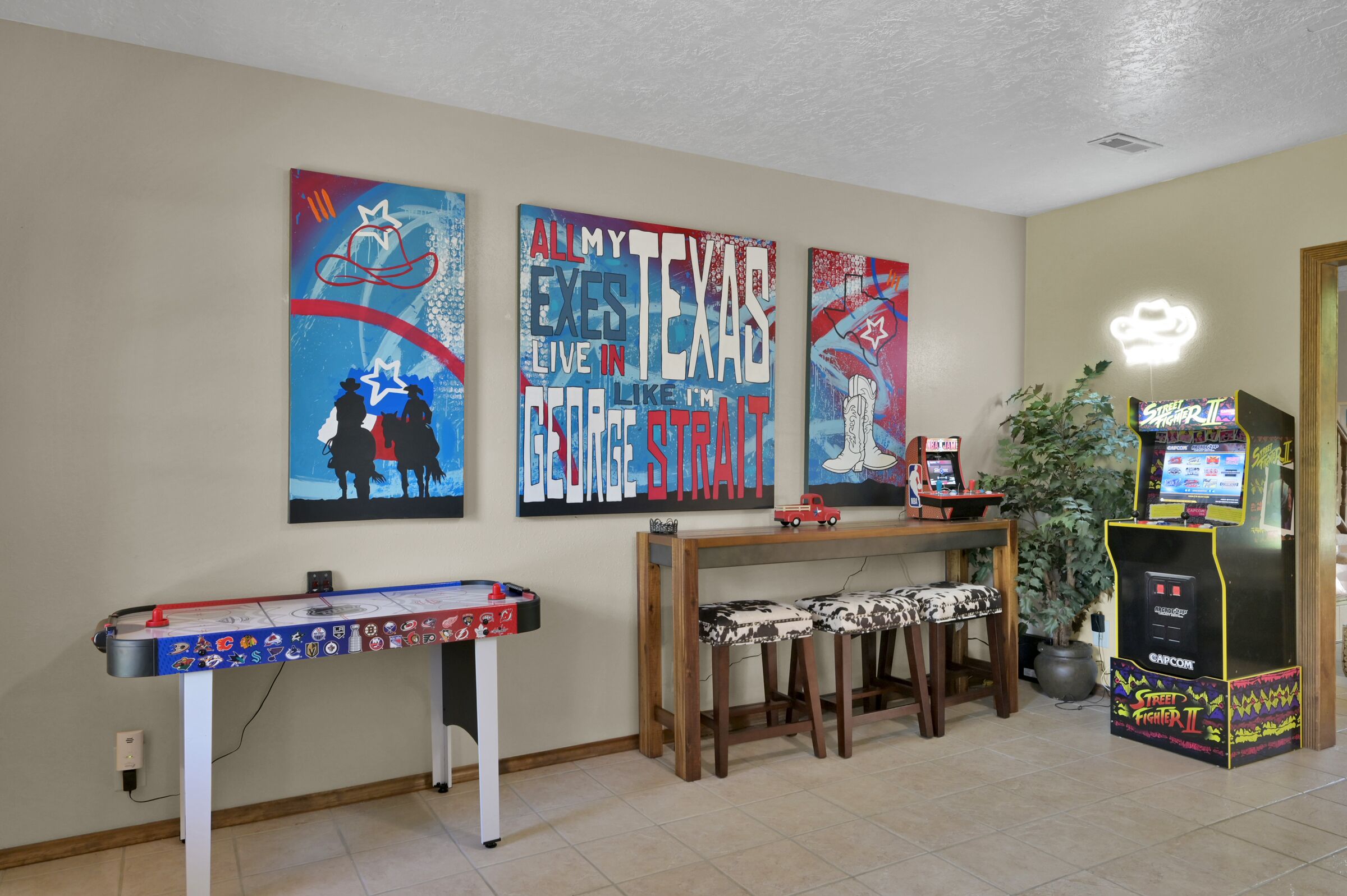 A room with a pool table, two wall art pieces, a console table with stools, and a video game machine.