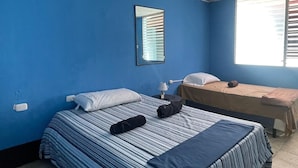 A room with two beds, one with striped bedding and the other with plain bedding. There is a window with blinds, a ceiling fan, and a wall-mounted mirror. - Hostal Cisne House (León)