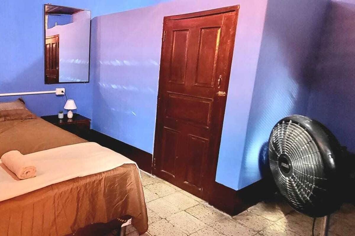 Ein Schlafzimmer mit einem Bett, einem Nachttisch, einem Spiegel, einer Tür und einem Ventilator.