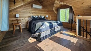 A cozy attic bedroom with a wooden sloped ceiling, a bed with a patterned bedspread, a small bedside table, and a window with a view of greenery.