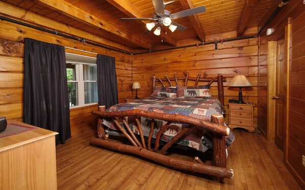 Un dormitorio rústico de cabaña de madera con una cama que tiene un cabecero de tronco, un ventilador de techo, una mesita de noche con lámpara y una ventana con cortinas.
