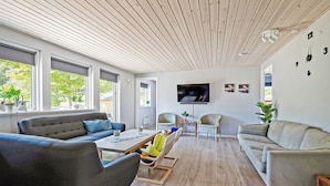 A modern living room with a wooden ceiling, large windows, a flat-screen TV, and a dining area with chairs and a table.
