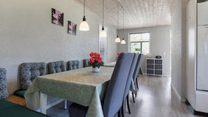 A dining area with a long table, chairs, pendant lights, and a refrigerator in the background.