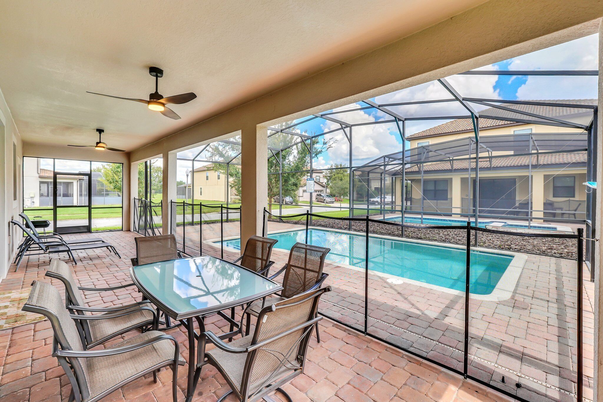 A covered patio with a glass table and chairs, overlooking a swimming pool and a fenced backyard.