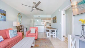 A living room with a pink sofa, a white coffee table, and a ceiling fan. There is a kitchen in the background with white cabinets and a dining table. - Island Winds West 871 2 Bedroom Condo (Gulf Shores)