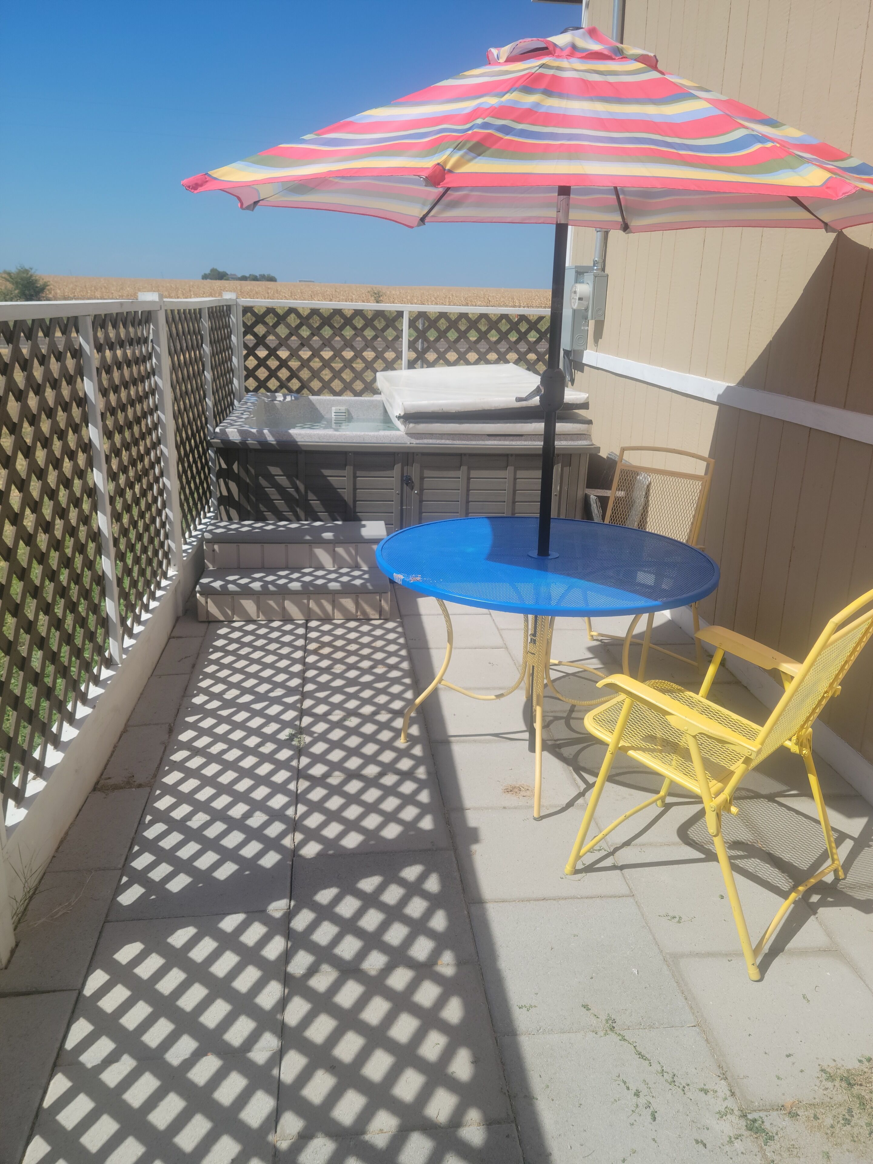 Un patio con un tavolo blu e sedie gialle, un ombrellone a righe e un recinto bianco.