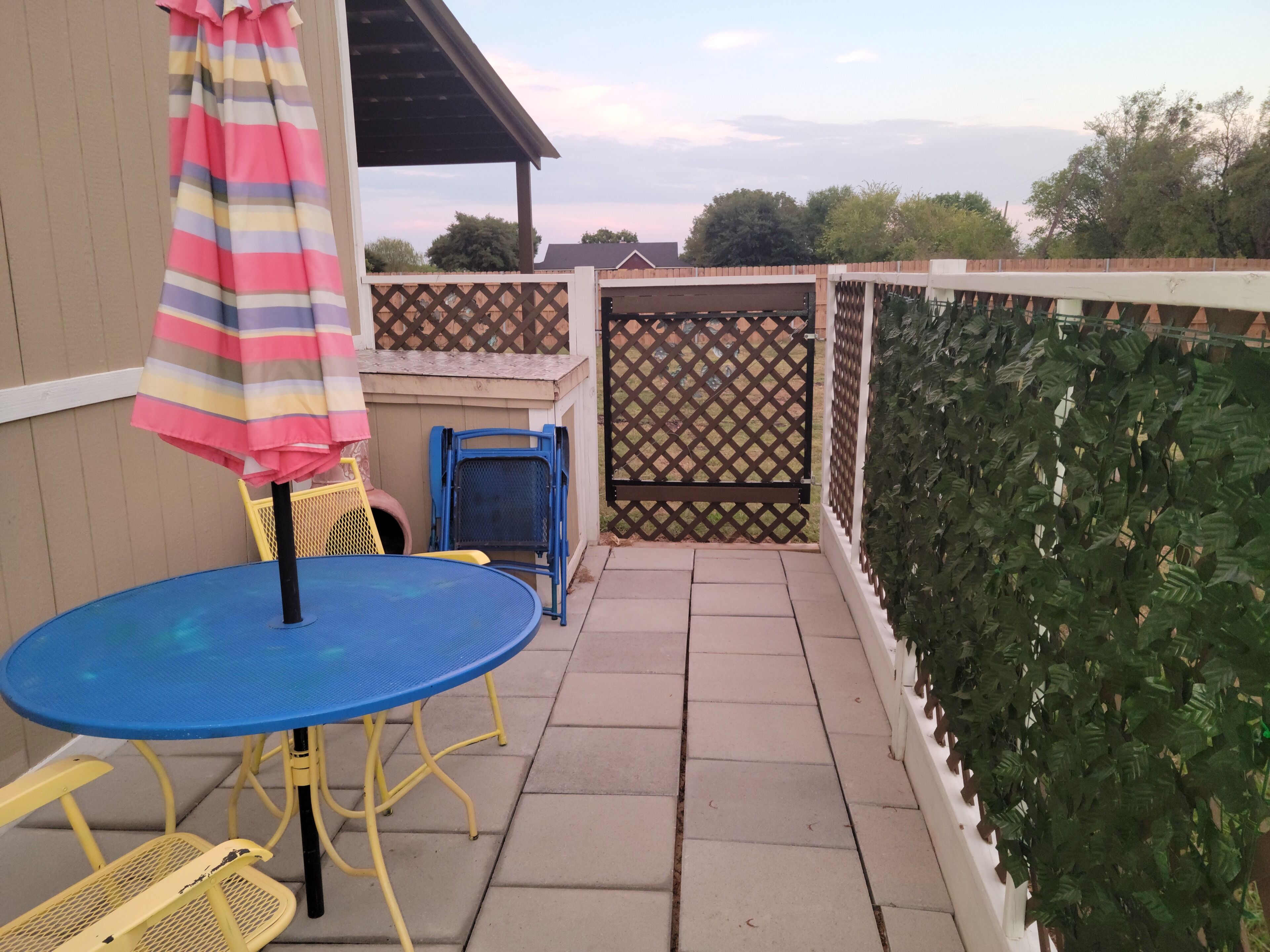 Une terrasse avec une table bleue et des chaises jaunes, un parasol à rayures et une clôture en bois.