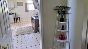 A hallway with a carpeted floor, a white door, a white shelf with plants, and a kitchen area in the background. - Kingdom Home Country Cottage is where you can relax, rejuvenate and enjoy peace. (Waxahachie)