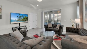 A modern living room with grey sofas, a glass coffee table, and a large flat-screen TV displaying a beach scene. - The Force At Solterra By Shine Villas #041 6 Bedroom Villa (Davenport)