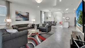 A modern living room with a grey sectional sofa, a glass coffee table, and a chandelier. There is a flat-screen TV mounted on the wall and a painting above the sofa. - The Force At Solterra By Shine Villas #041 6 Bedroom Villa (Davenport)