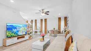 Una sala de estar moderna con una televisión de pantalla plana, un sofá blanco, una mesa de centro con flores, un área de comedor con una mesa y sillas, y un ventilador de techo.