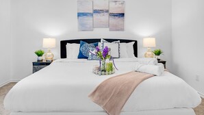 Un dormitorio ordenado con una cama grande, dos mesitas de noche con lámparas, un jarrón decorativo con flores y una obra de arte colgada en la pared sobre la cama.