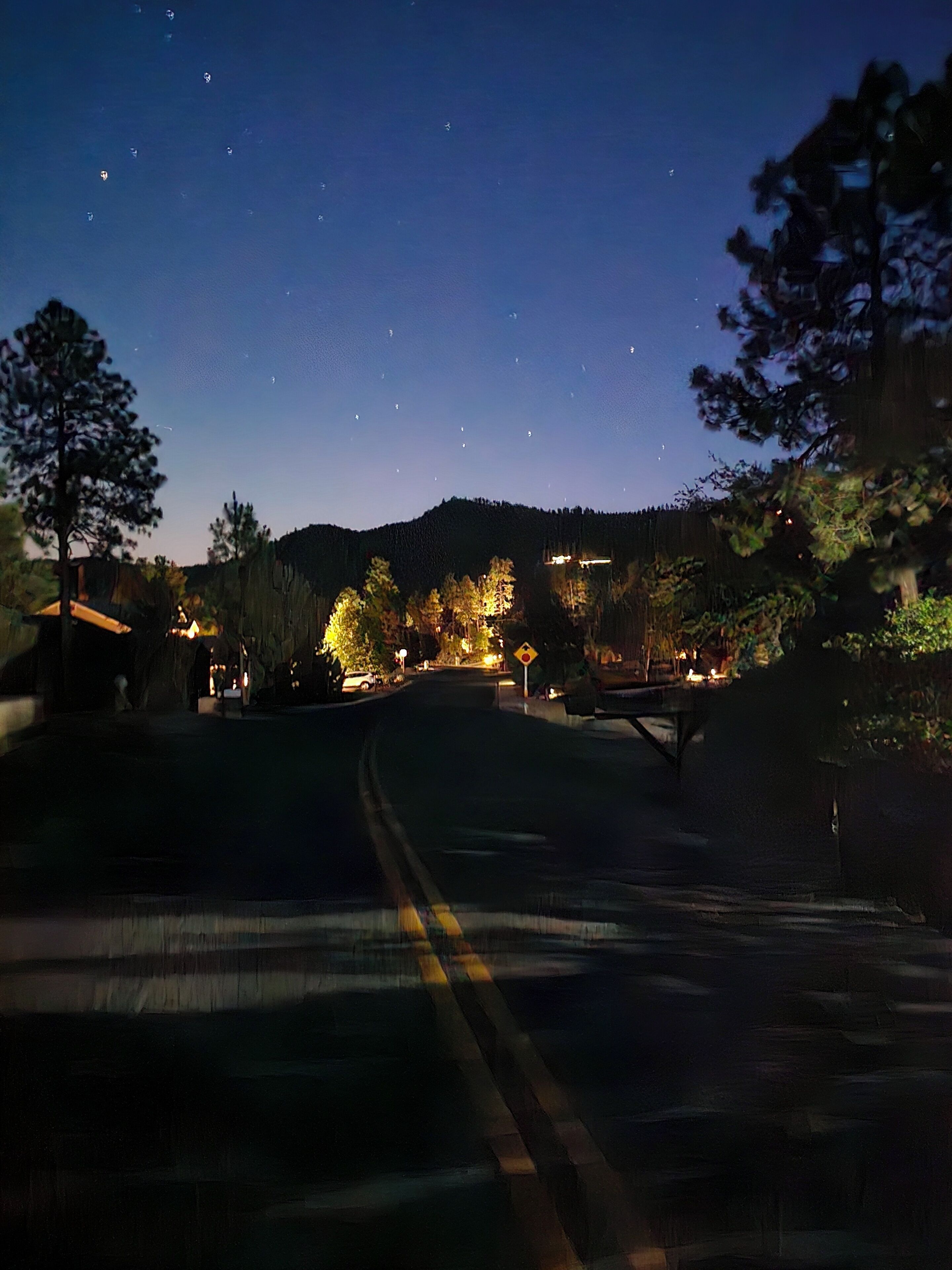 A nighttime street view with a road leading to a residential area, houses with illuminated windows, and a hill in the background.