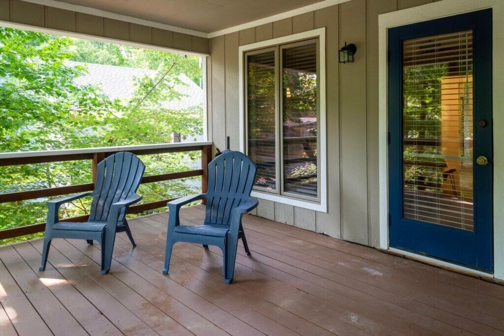 Un porche con dos sillas Adirondack, una terraza de madera, una puerta azul con un ventanal y vistas a la vegetación.