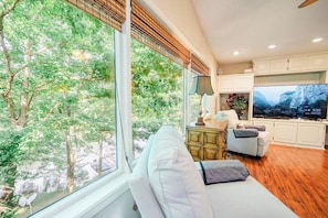 A living room with a sofa, a television, and a view of trees through large windows. - Lakeside Day Dreamin (Hot Springs Village)