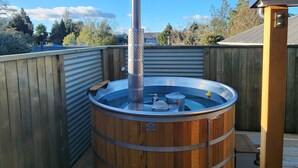 Ein Whirlpool auf einer Holzterrasse mit einem Metallschornstein, umgeben von einem Holzzaun und blauem Himmel.