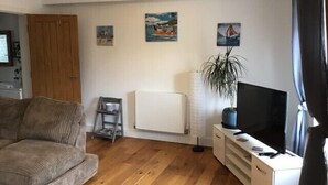 A living room with a wooden floor, a grey sofa, a white radiator, a TV on a white stand, and a small plant. - Central Teignmouth Penthouse, Sleeps 4, Pets Ok (Teignmouth)