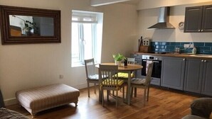 A cozy living room with a wooden floor, a round dining table with chairs, a bench, and a kitchen area with grey cabinets and a stove. - Central Teignmouth Penthouse, Sleeps 4, Pets Ok (Teignmouth)
