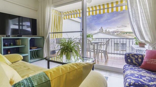 Ein Wohnzimmer mit Sofa, Fernseher und einem Balkon mit gelb-weiß gestreiften Markisen.