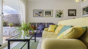 Ein Wohnzimmer mit einer Couch, einem Couchtisch und einem Blumentopf. An der Wand hängen Bilder und es gibt ein Fenster mit Vorhängen.