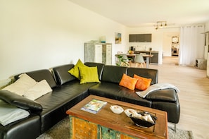 Ein modernes Wohnzimmer mit einem schwarzen Ledersofa, einem Holztisch und einer Küche im Hintergrund.