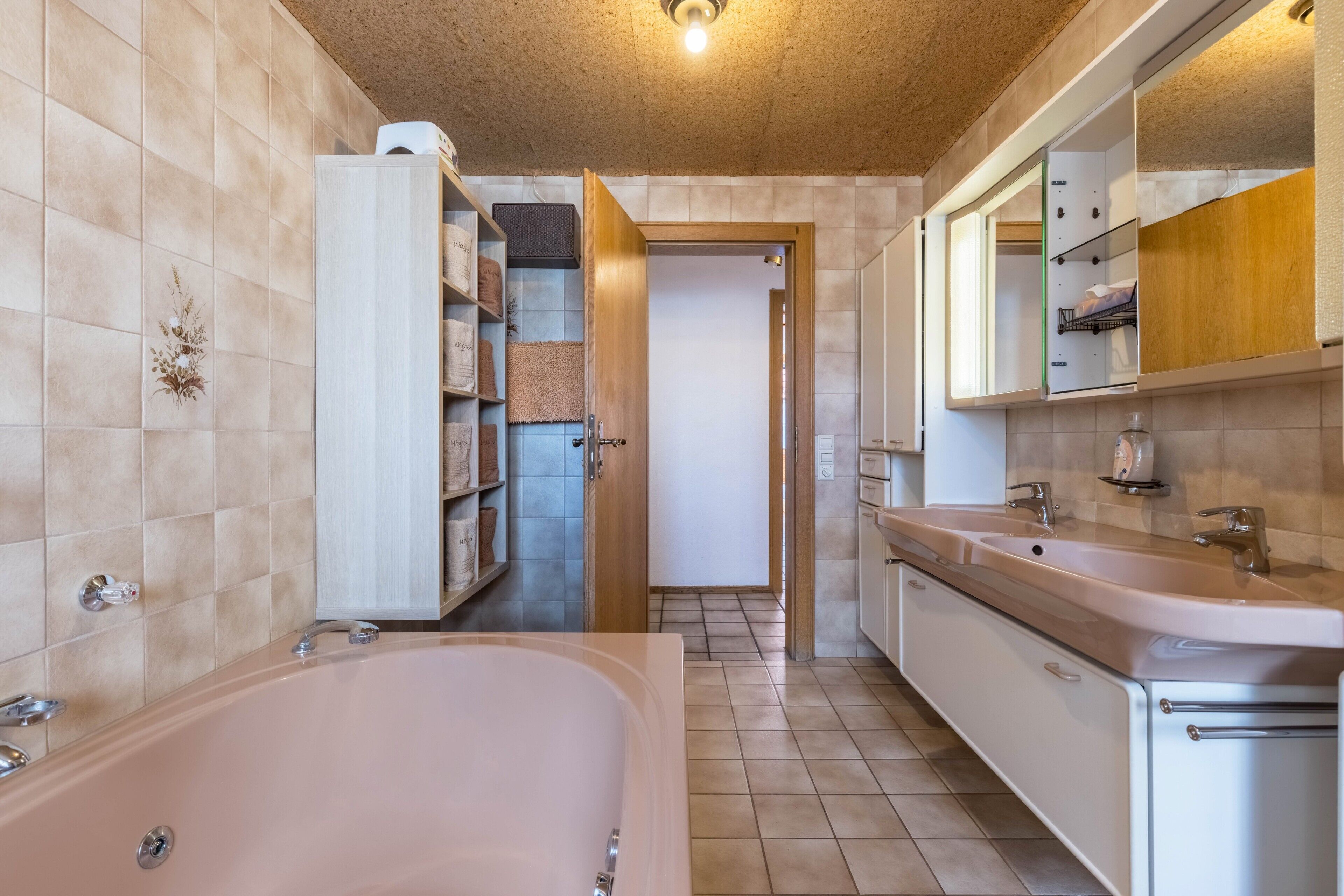 A bathroom with a bathtub, a sink, and a mirror. There is a shelf with towels and a door leading to another room.
