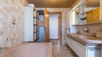 A bathroom with a bathtub, a sink, and a mirror. There is a shelf with towels and a door leading to another room.