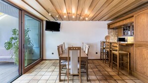 A dining area with a wooden table and chairs, a television mounted on the wall, and a kitchen area with wooden cabinets.