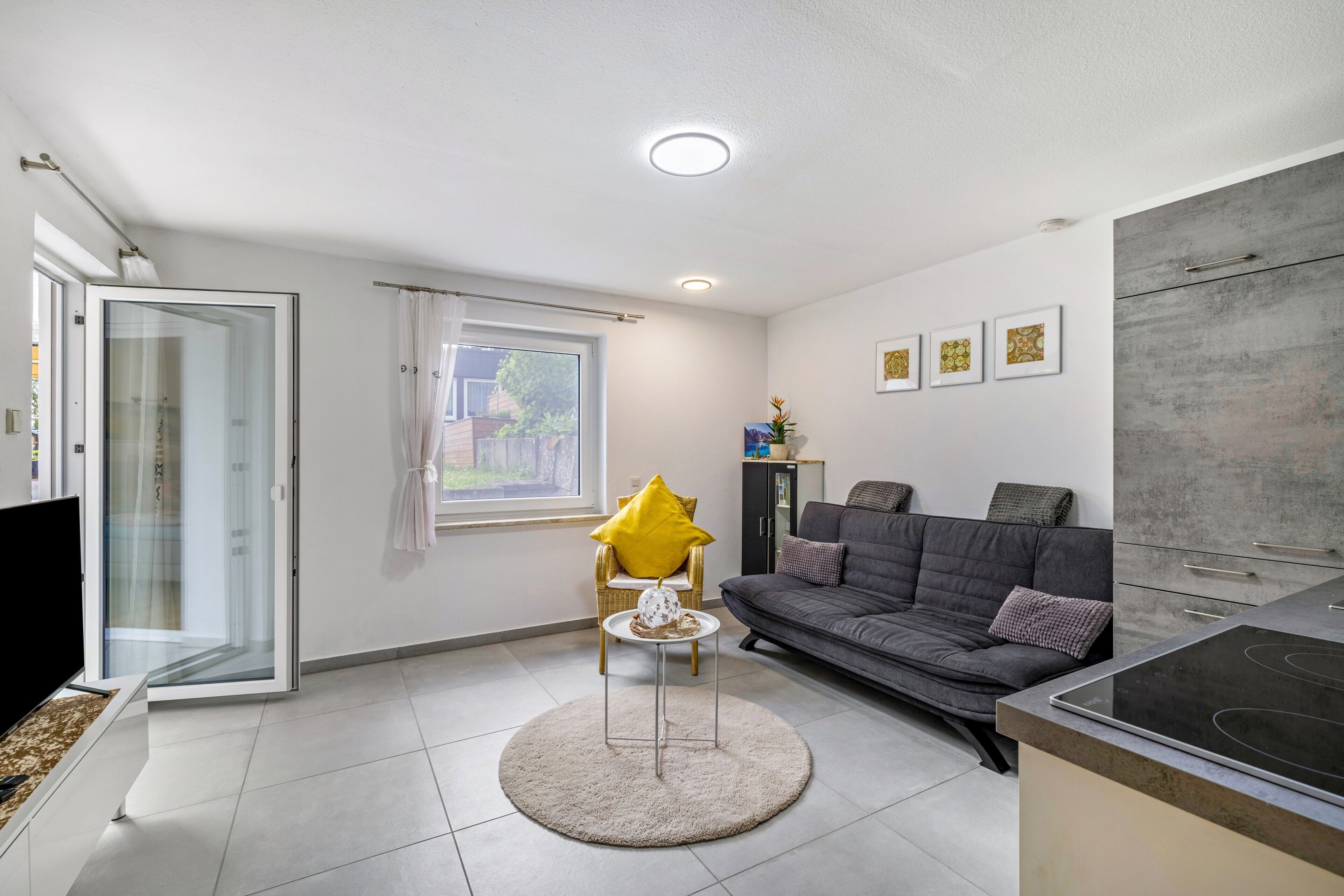 A modern living room with a sofa, a small round table with a yellow cushion, a glass door leading outside, and a kitchen area with a stove.