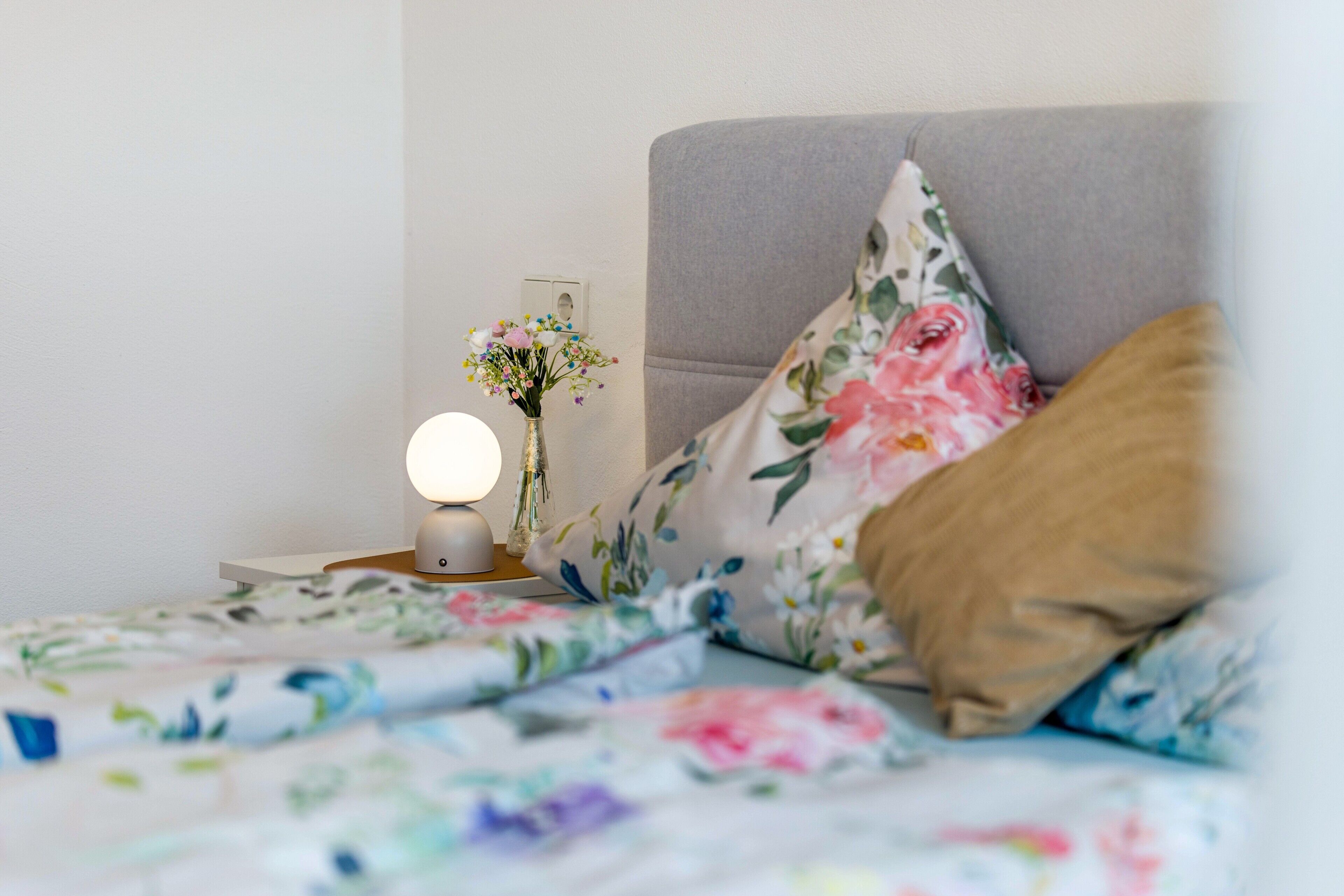 A bed with floral bedding, a bedside lamp, and a vase with flowers.