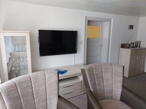 A room with a flat-screen TV, a desk with drawers, two patterned chairs, a glass cabinet with wine glasses, and a door with a patterned panel.