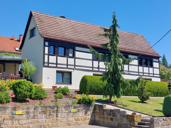 A two-story house with a sloped roof, a stone wall with a parking sign, and well-maintained gardens.