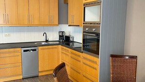 Cocina compacta con armarios de madera, lavavajillas, cafetera y comedor con sillas de mimbre y mesa de madera.