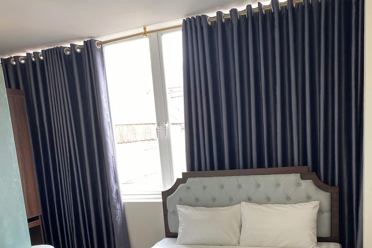 A neatly made bed with white linens, a wooden headboard, and a window with dark curtains.