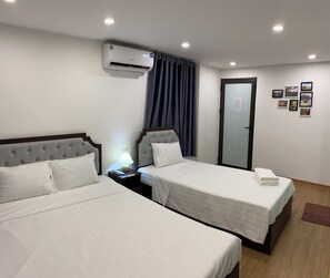 Une chambre d’hôtel avec deux lits, un climatiseur, une table de chevet avec une lampe, une porte avec un panneau en verre et des tableaux encadrés au mur.