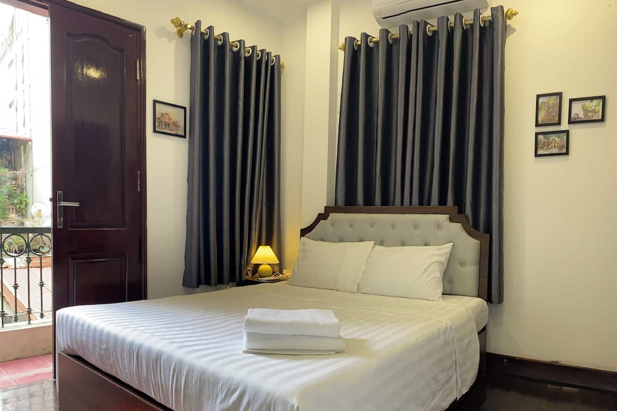 A hotel room with a bed, curtains, a nightstand with a lamp, and a door.