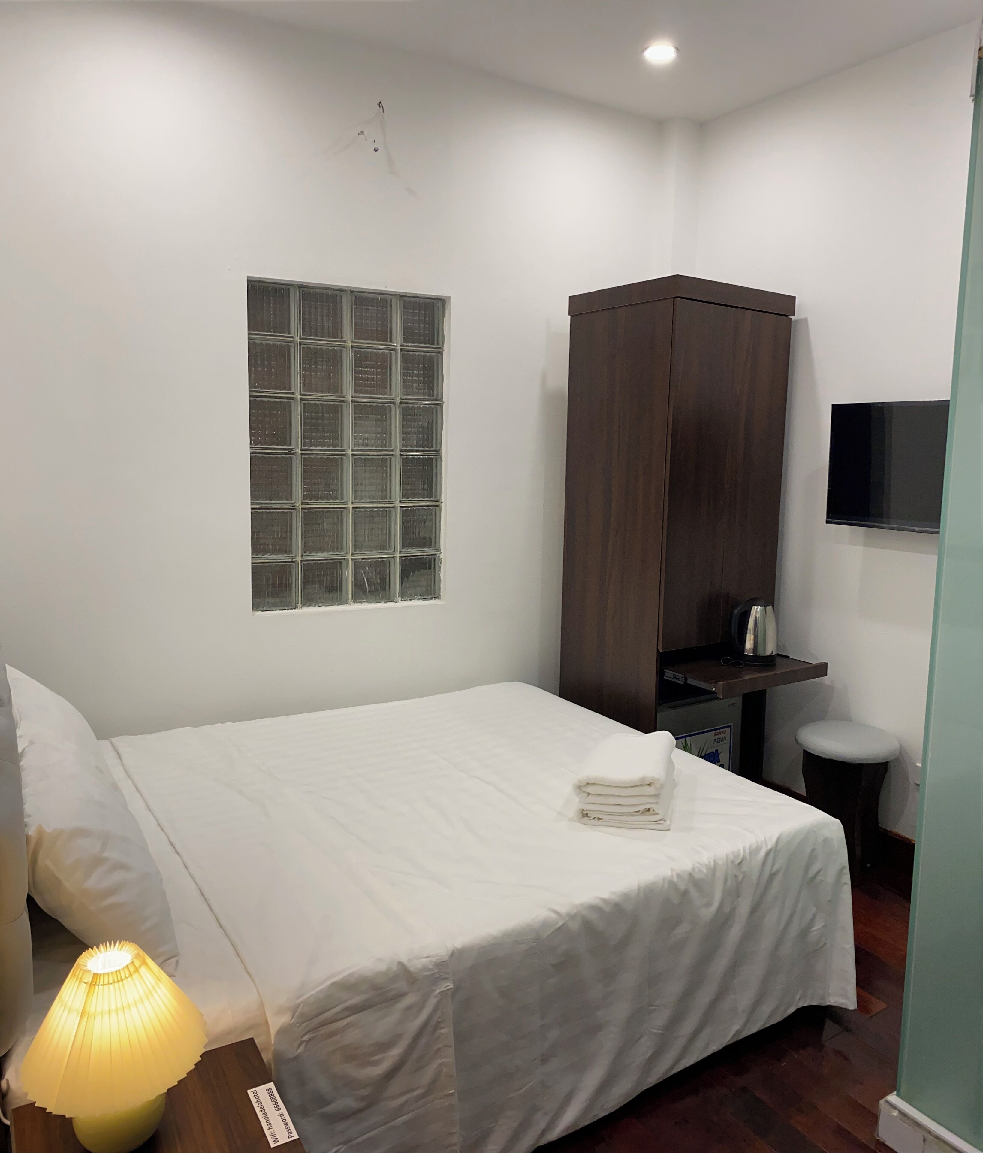 A neatly made bed with a white duvet, a wooden wardrobe, a small table with a kettle, and a wall-mounted TV.