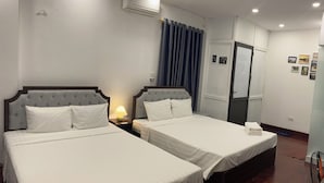 Een hotelkamer met twee bedden, een plafondventilator, een airconditioningapparaat, een nachtkastje met een lamp, een deur en een muur met ingekaderde foto's.