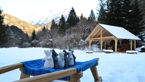 Une luge en bois avec une couverture bleue et violette, un chalet en bois dont le toit est recouvert de neige, et des arbres enneigés en arrière-plan.