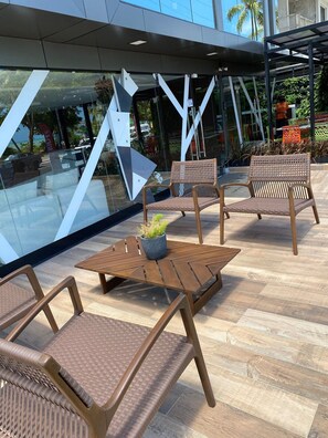 Un área de estar al aire libre con mobiliario de mimbre y una mesa de madera, con vistas a una piscina y un edificio moderno con estructuras de vidrio y metal.