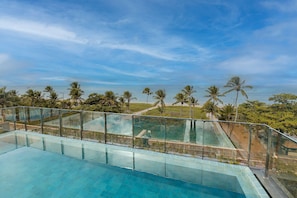 A clear view of a swimming pool with a glass barrier, surrounded by palm trees and a beach in the background. - Fortune Cabo Branco (João Pessoa)