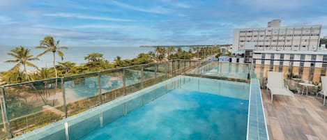 A rooftop infinity pool with a glass barrier, overlooking a beach and palm trees.