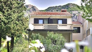 Una casa de dos pisos con una antena parabólica, un balcón con barandilla de celosía y un paisaje montañoso de fondo.