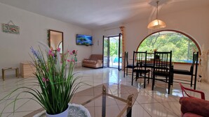 Amplia sala de estar con sofá, mesa de comedor, sillas, una planta en maceta y un ventanal con vistas a la vegetación.