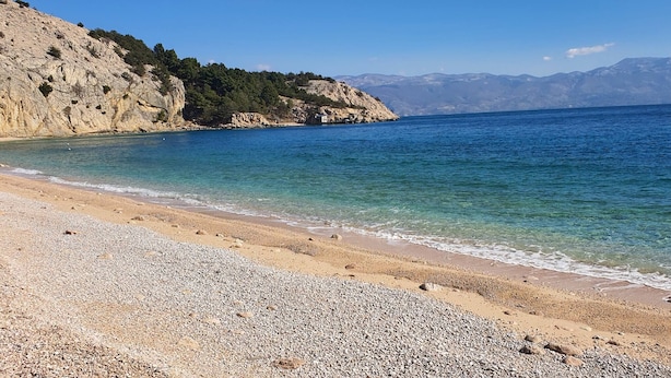 Una playa con agua cristalina, orilla pedregosa y un acantilado rocoso.